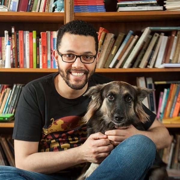 Tônio Caetano sentado em frente a uma estante de livros abraçando sua cachorrinha Safira, de pelos pretos e marrons