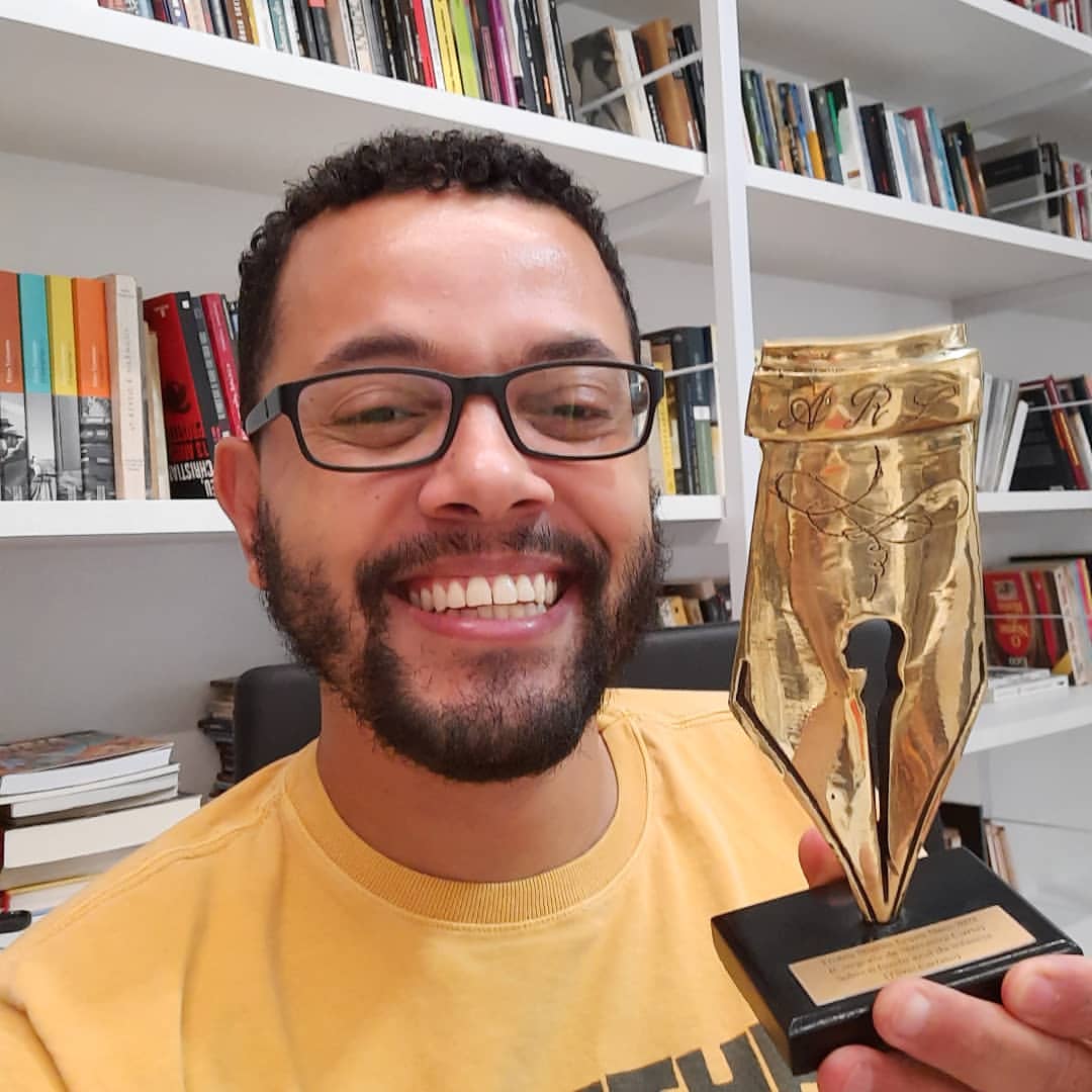 Retrato de Tônio Caetano em sua casa vibrando com troféu do Prêmio da Academia Rio-Grandense de Letras de 2021